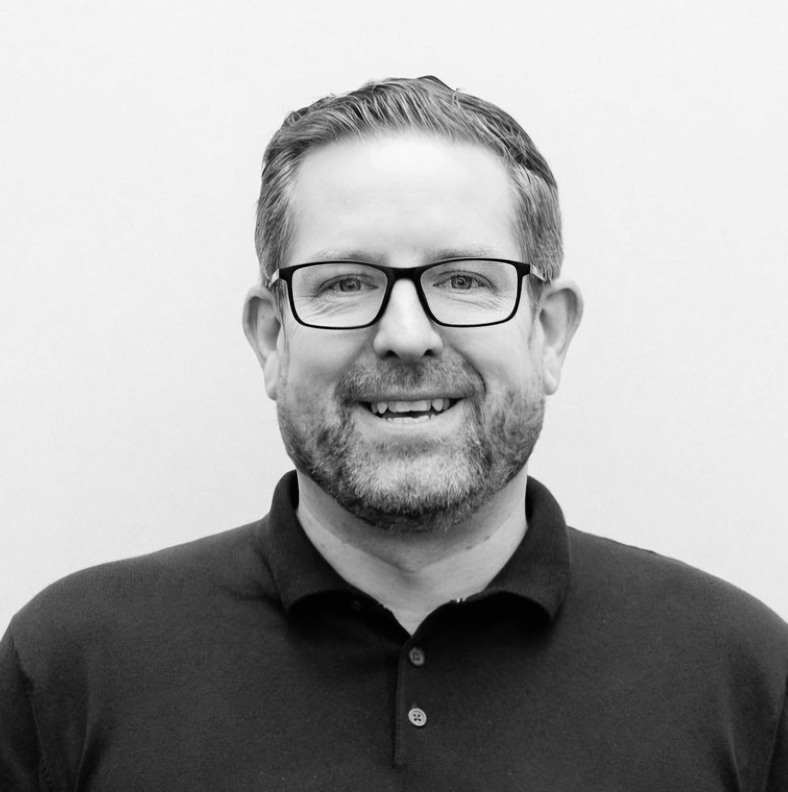 a black and white image of a smiling man in a polo shirt with a beard and glasses.